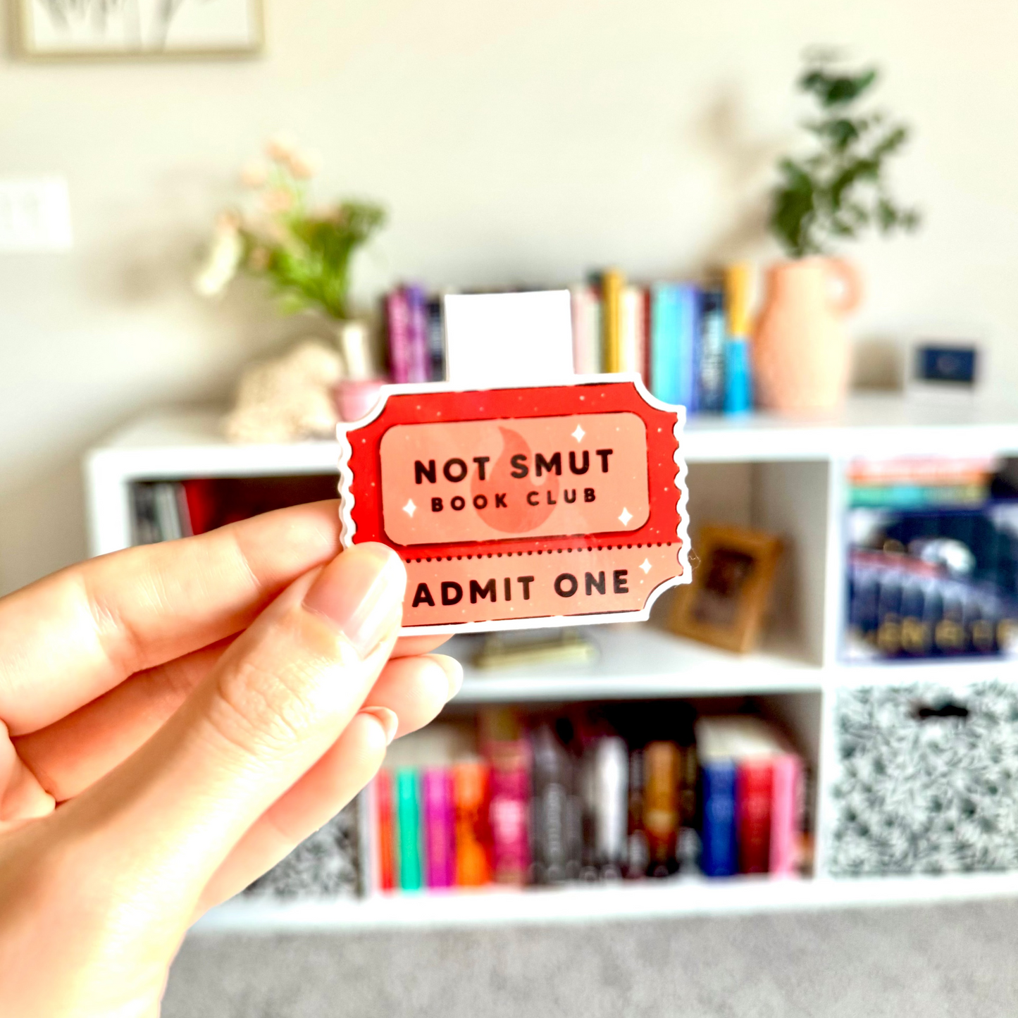 Hand holding a 'Not Smut Book Club' admit one ticket with a bookshelf in the background.